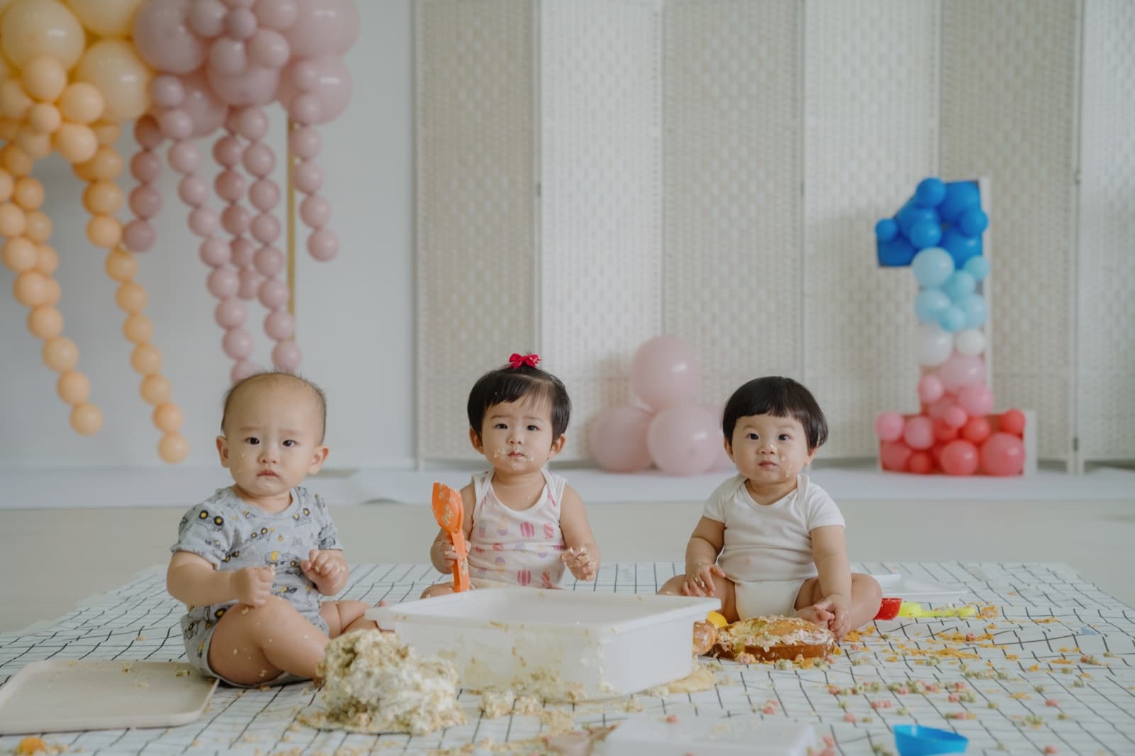 Three babies enjoying a first-birthday cake-smash surrounded by pink and peach balloon garlands